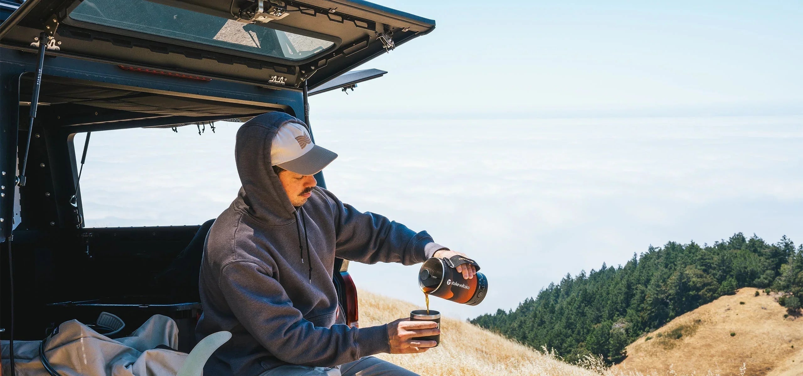 Camper pours coffee from Stoke Voltaics kettle pot outdoors by SUV with mountain view