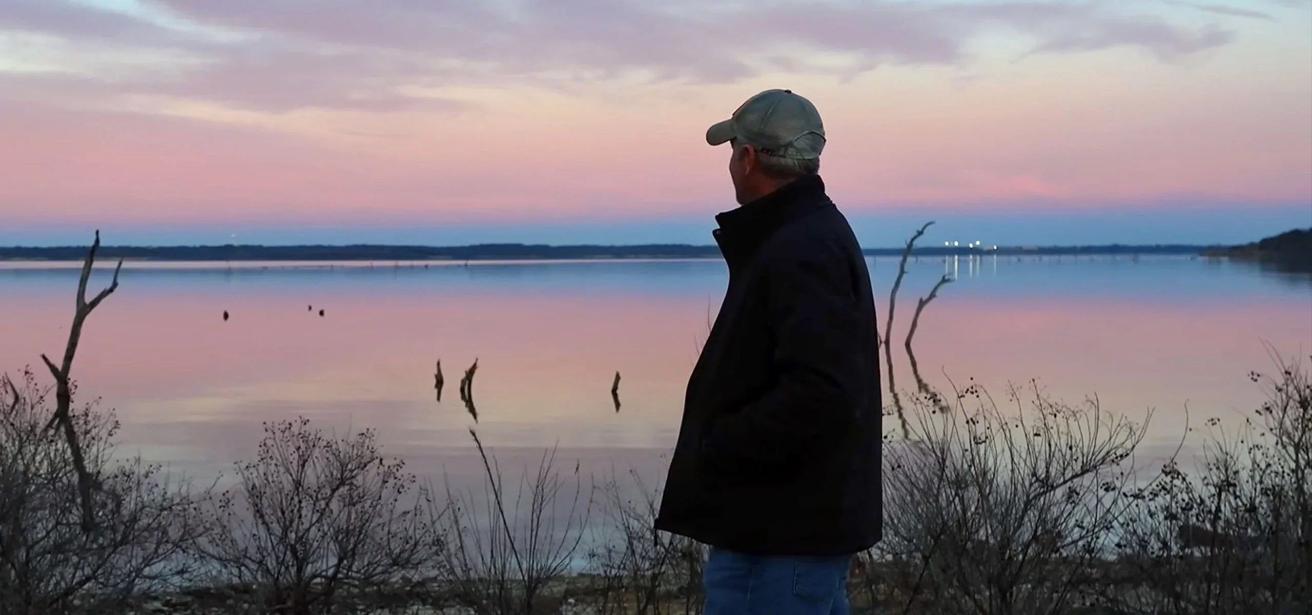 Man in cap and jacket looking at serene lake during colorful sunset, nature outdoors