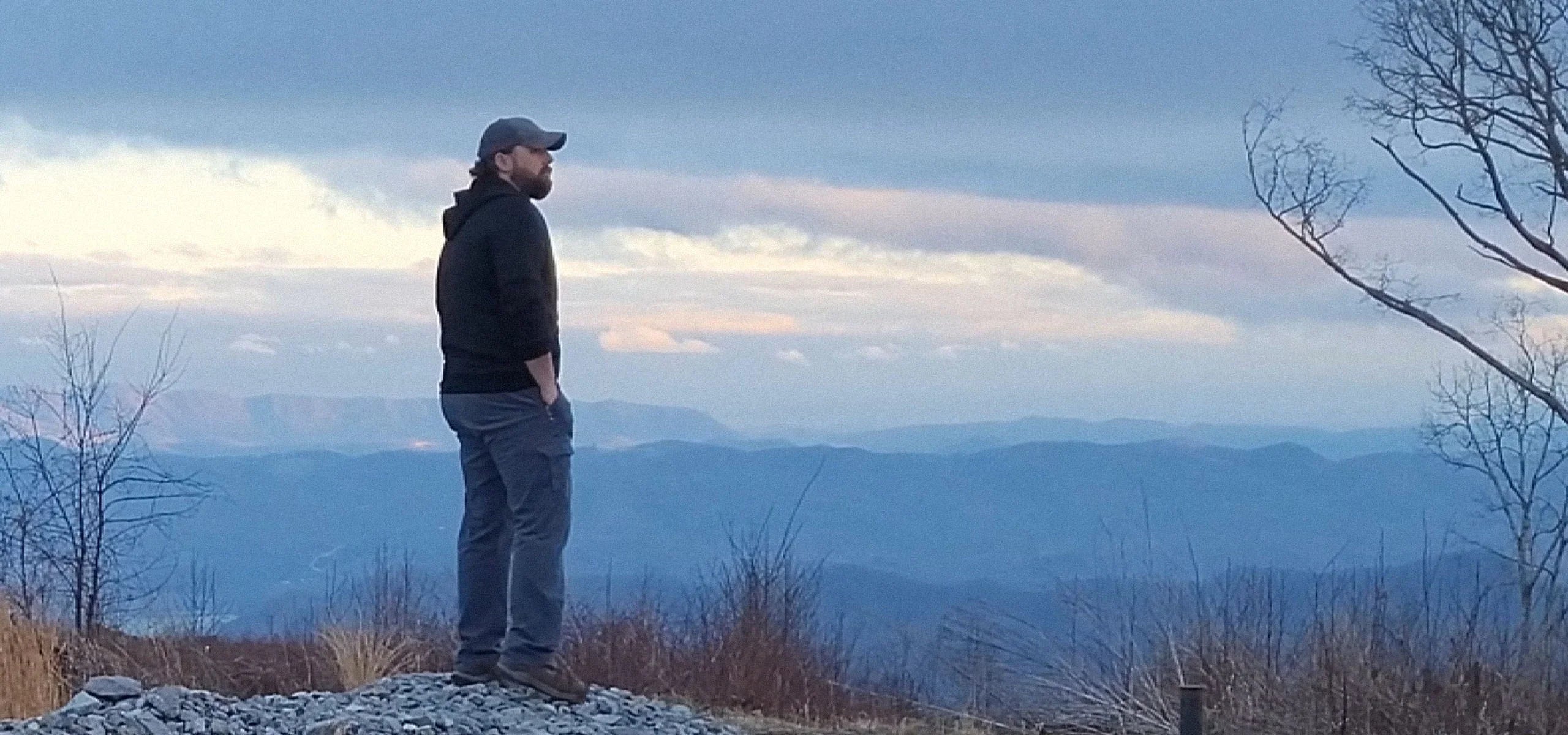 Man in outdoor gear stands on rocky hilltop overlooking scenic mountain landscape at sunset