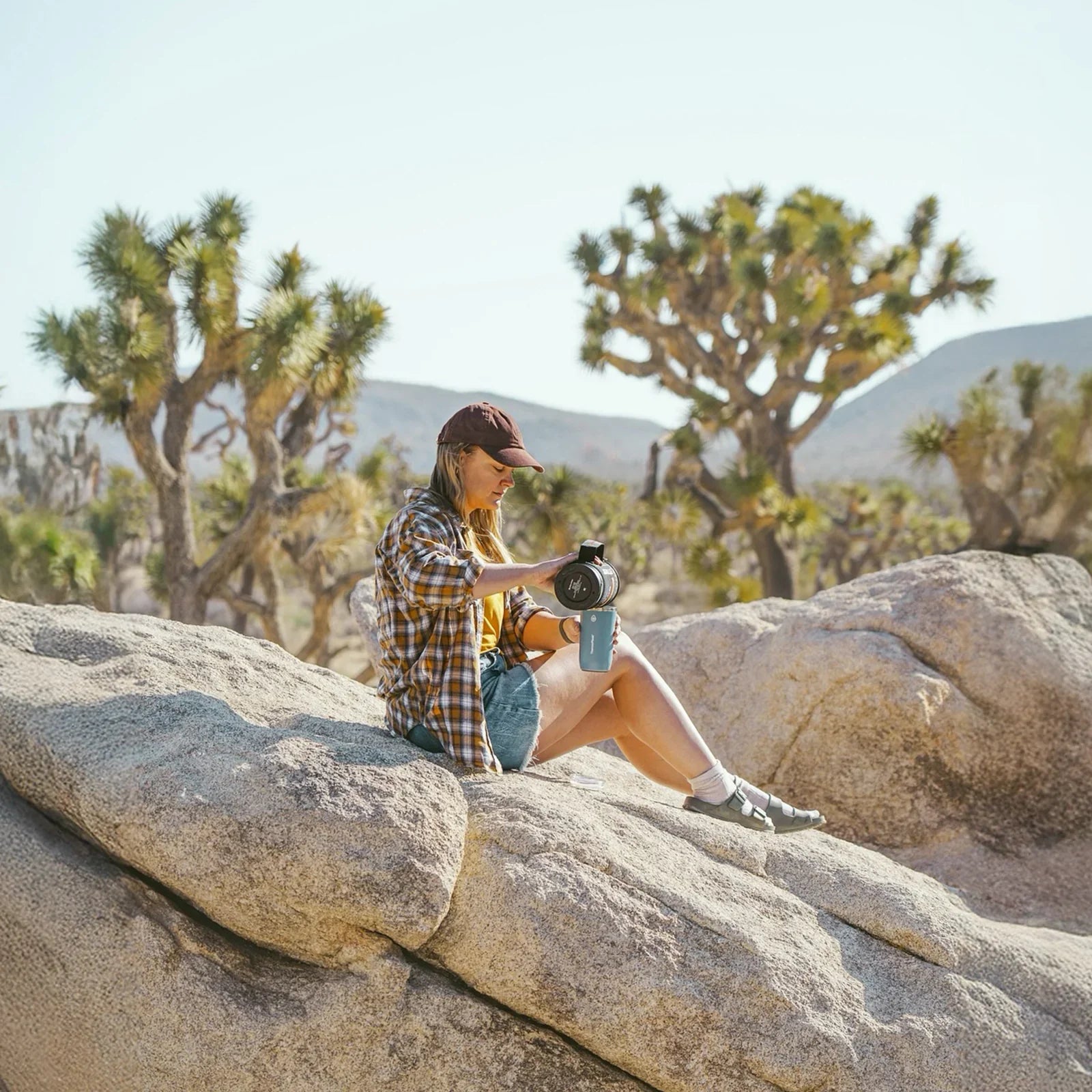 Woman using outdoor coffee maker on rocks in desert with Joshua trees, Stoke Voltaics camping gear